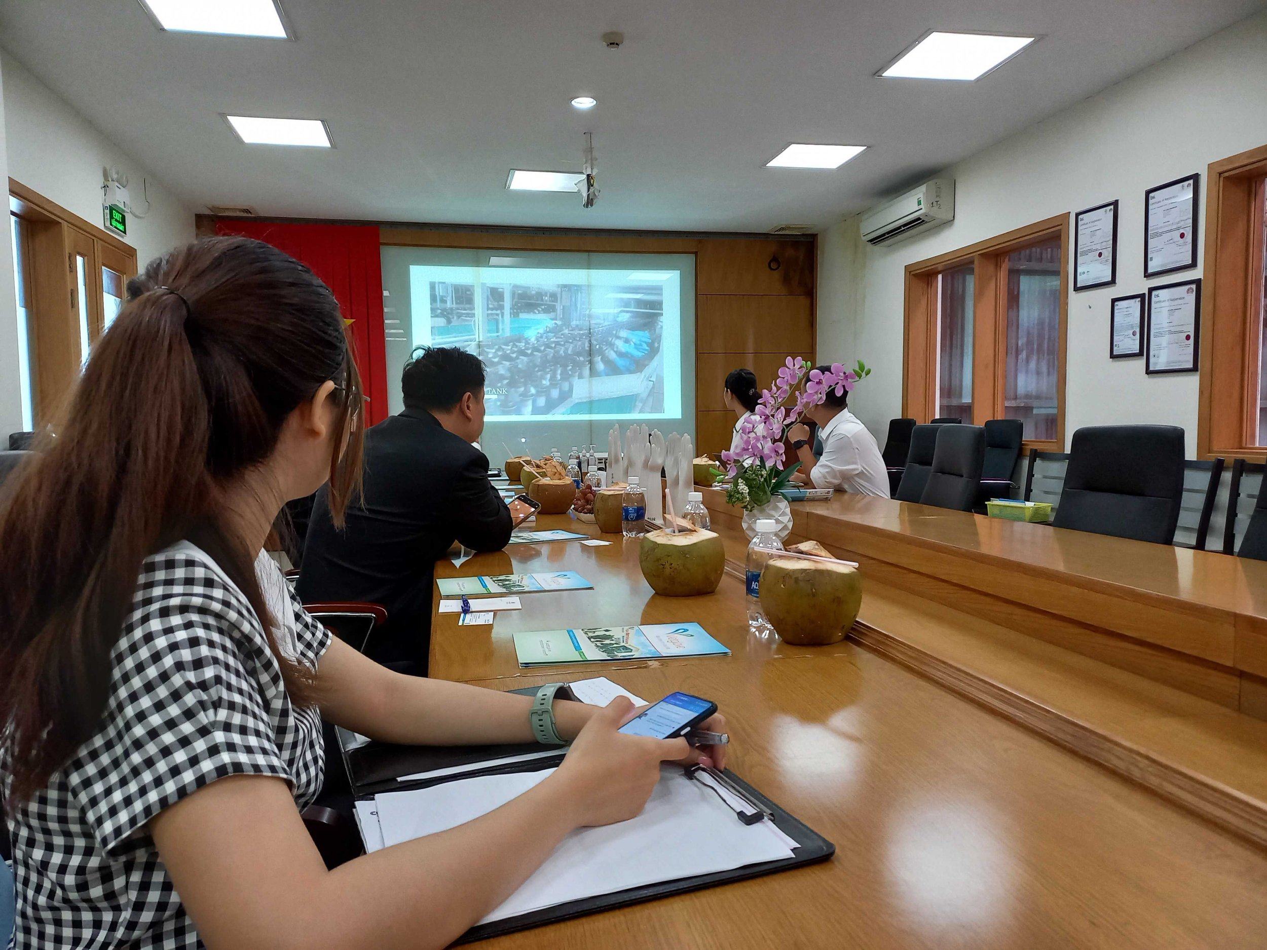 Four people sitting around a conference table in a meeting room, some using phones or looking forward. There is a projector screen, bottled water, notebooks, and decorative vases with pink flowers on the table. The room has wooden-framed windows and framed certificates on the wall.