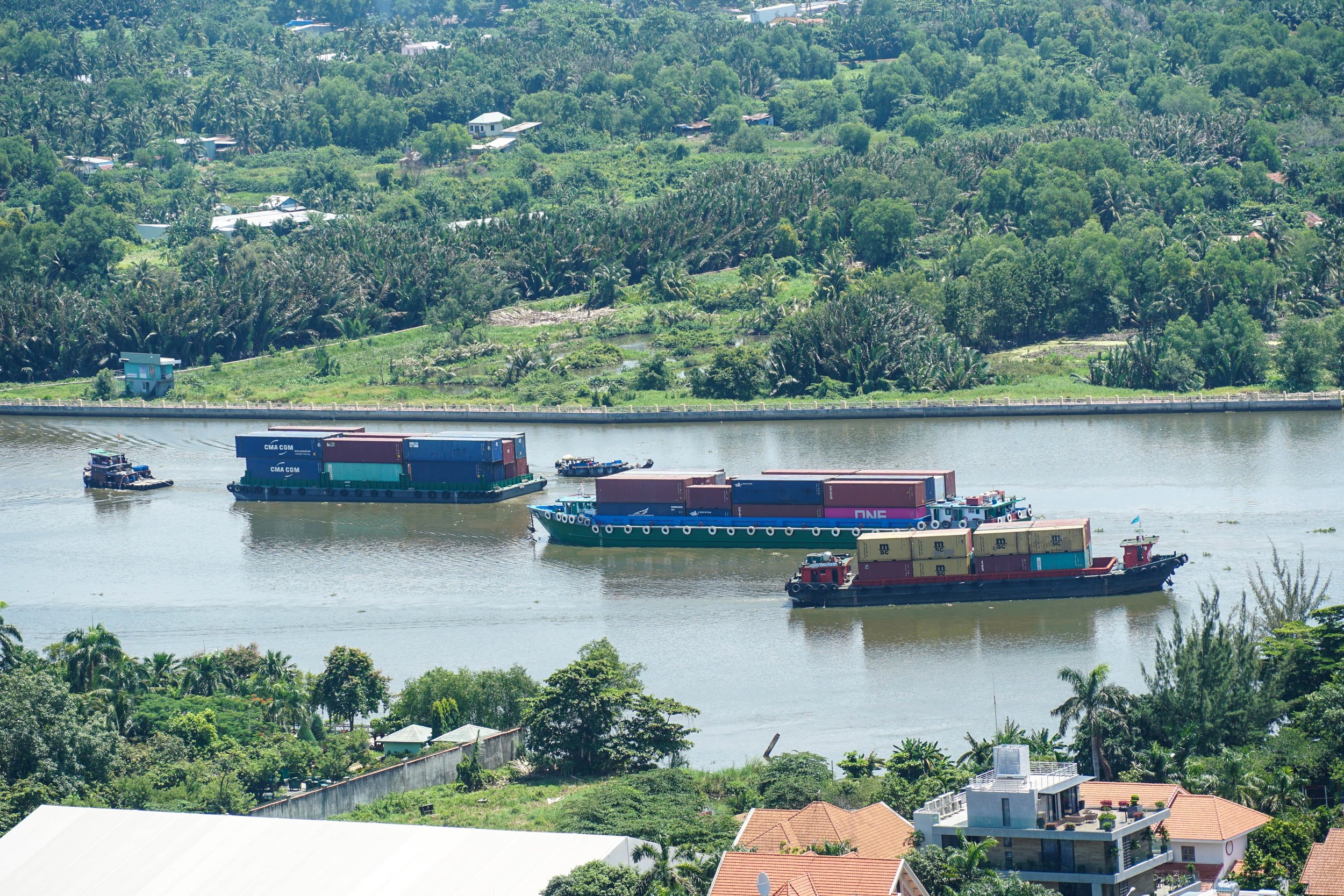 Three cargo boats carrying containers travel along a river with lush green trees and houses in the background.