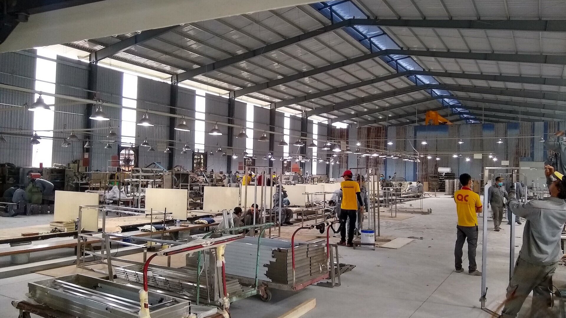 Inside a spacious factory with workers wearing masks, some in yellow shirts and red helmets, engaged in manufacturing activities. The area is well-lit with overhead lights and has industrial machinery and materials scattered throughout.